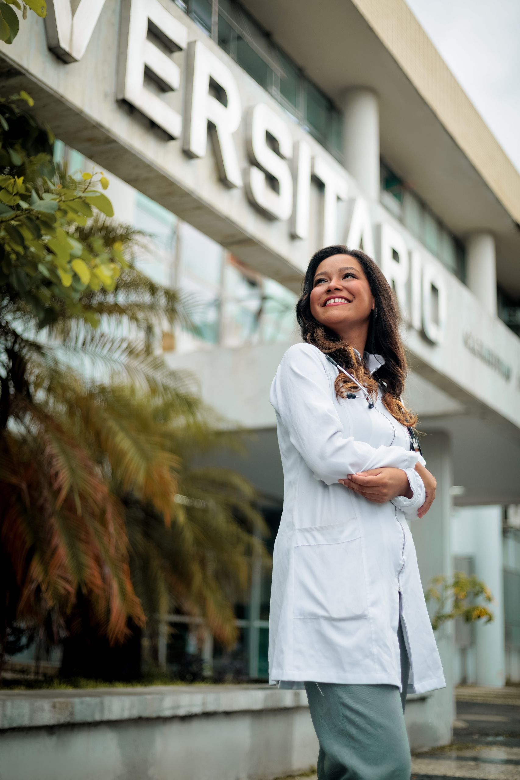 Smiling female doctor in white coat standing outdoors near university building, expressing confidence and optimism.
